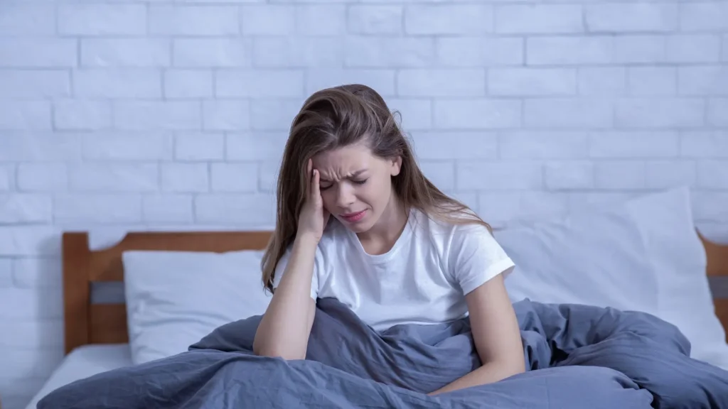 A woman sitting on a bed with her head in her hands, looking distressed.