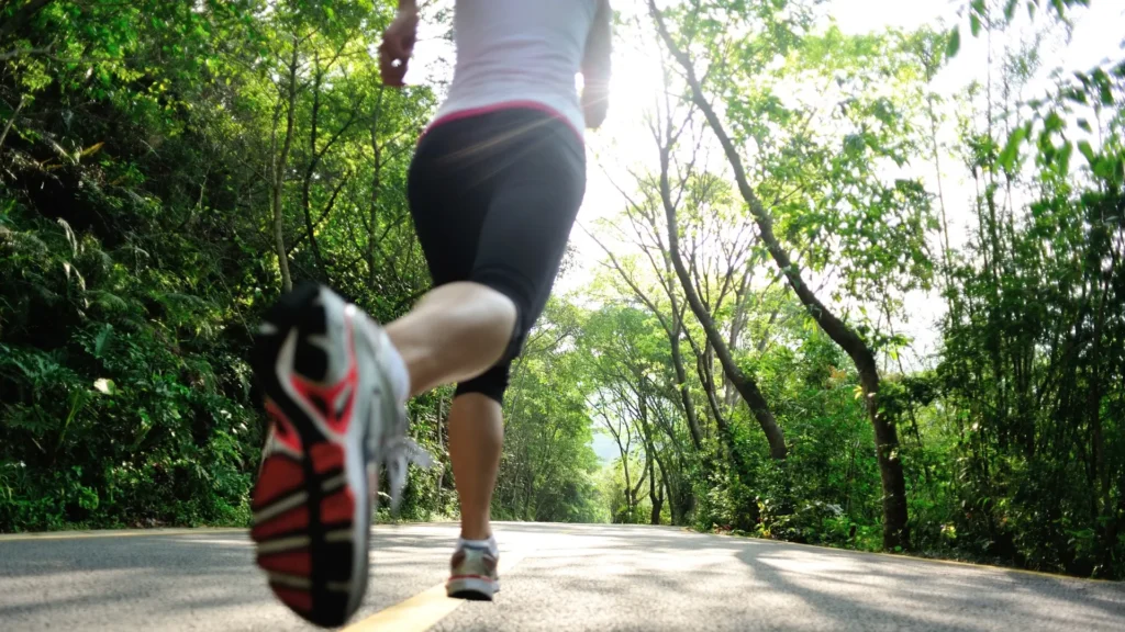 Woman jogging on forest road, surrounded by trees, under a clear sky.