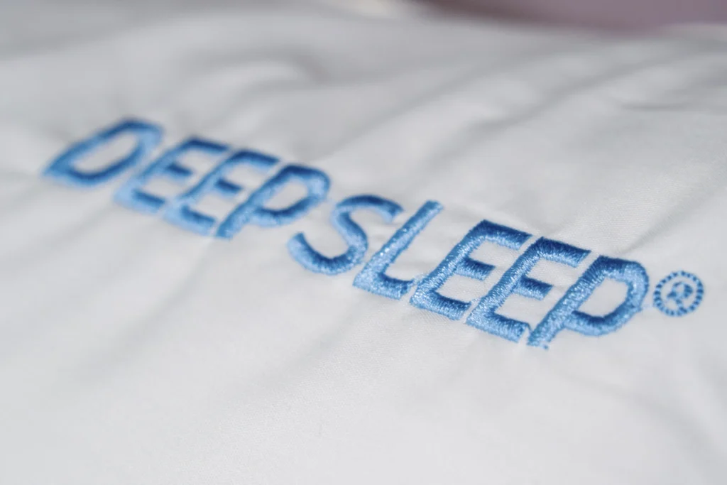 Close-up of the embroidered "DEEPSLEEP" logo in blue on a Simmons DeepSleep pillow, highlighting its premium branding and soft fabric.