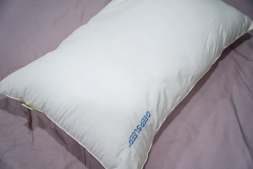 A Simmons DeepSleep pillow placed on a lavender bedsheet, showcasing its sleek white design and embroidered "DEEPSLEEP" logo for a luxurious sleeping experience.